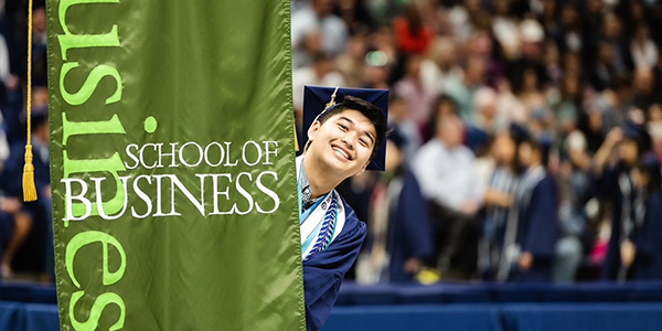 A student in a cap and gown smiling and posing with the School of Business banner at commencement.